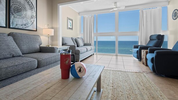 A living room with a grey sofa, a red thermos, and a beach ball on a wooden coffee table. There are blue armchairs and a white lamp. The room has large windows with a view of the ocean. - GULF-FRONT VIEW, POOL, HEART OF GULF SHORES! (Gulf Shores)