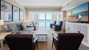 A spacious living room with a large window overlooking the ocean, a flat-screen TV, and a beach-themed painting. - GULF-FRONT VIEW, POOL, HEART OF GULF SHORES! (Gulf Shores)