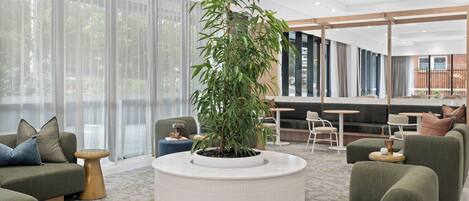 A modern living room with a large potted plant, green sofas, a round coffee table, and a dining area with a brick wall and large windows.