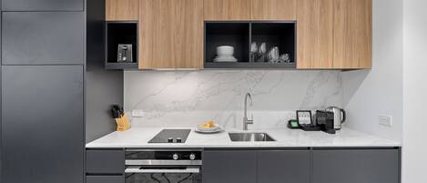 Una cocina moderna con armarios grises, un estante superior de madera, salpicadero de mármol y electrodomésticos integrados, incluyendo lavavajillas, horno y cafetera.