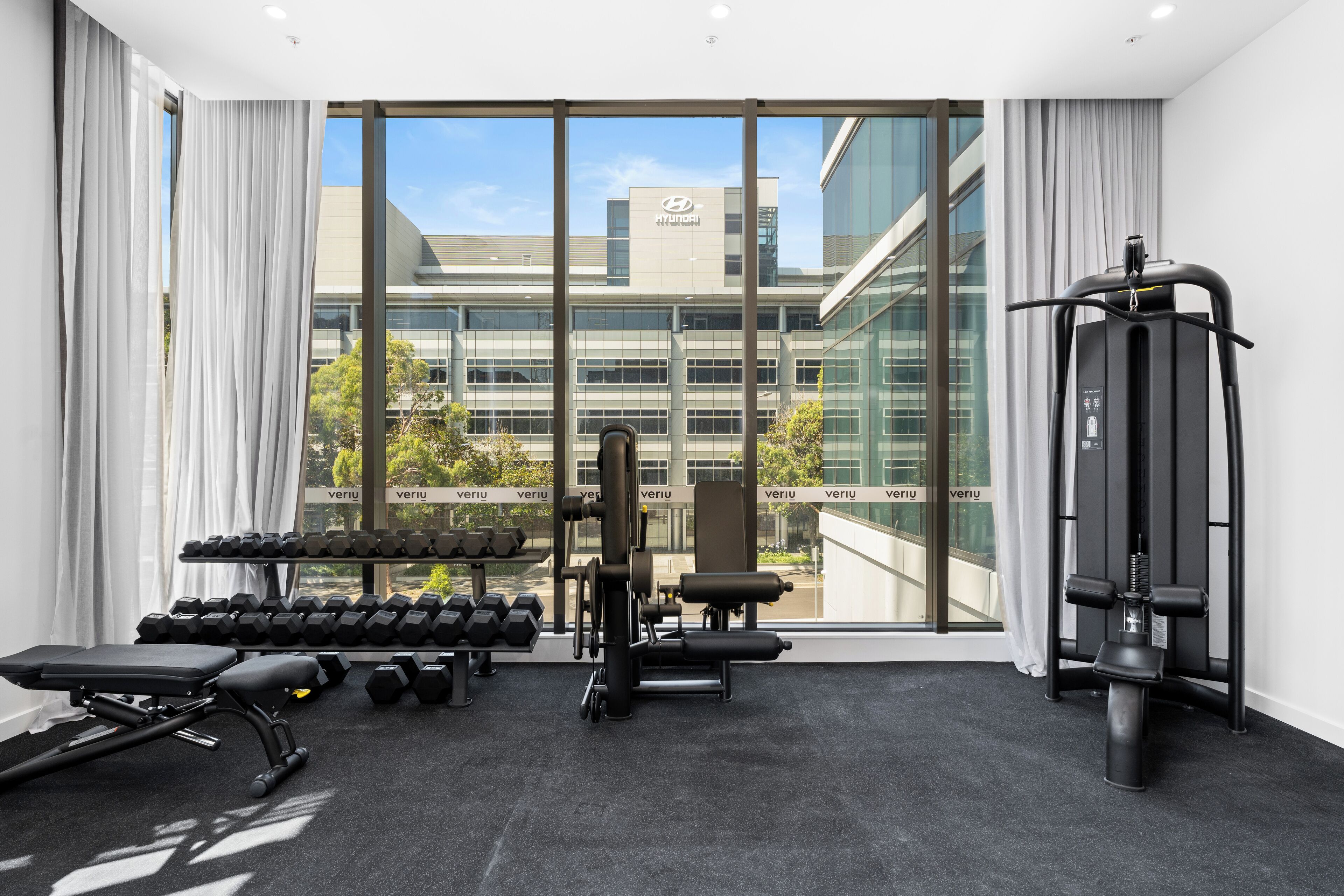 A gym with dumbbells, benches, and weight machines, large windows, and a view of a building with 'HYUNDAI' signage.