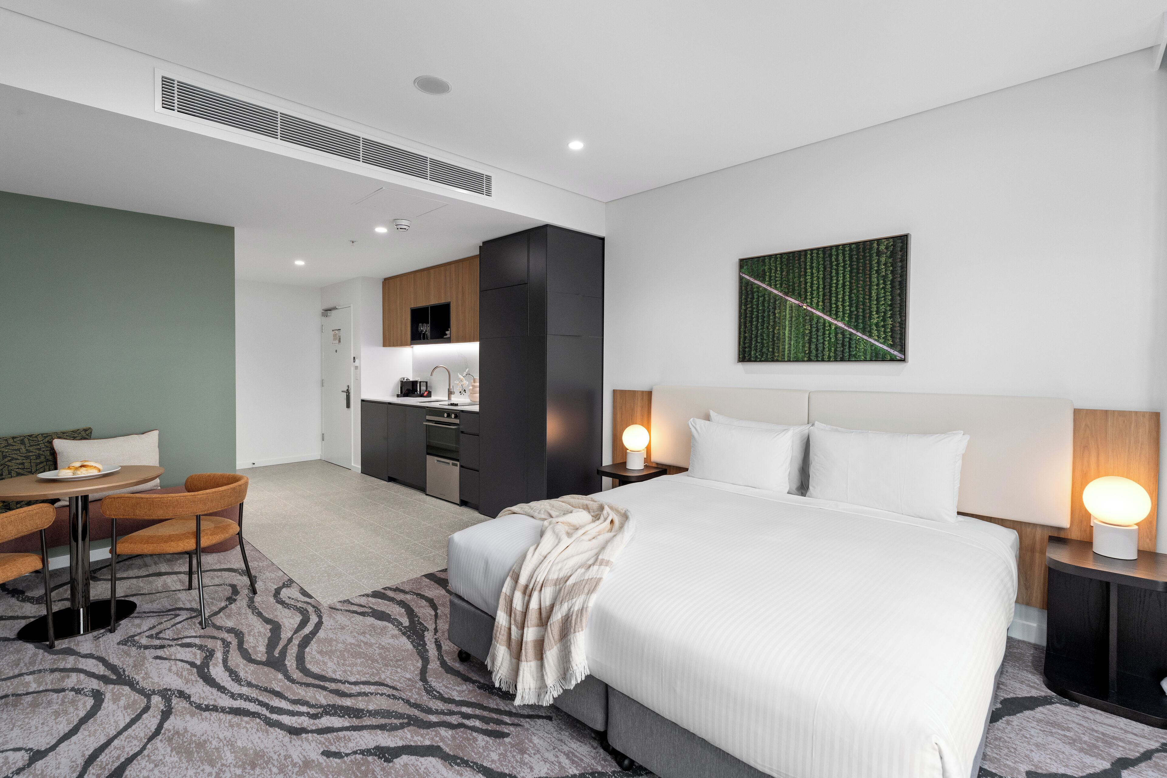 A modern hotel room with a large bed, a dining area with a table and chairs, a kitchenette, and a bathroom.