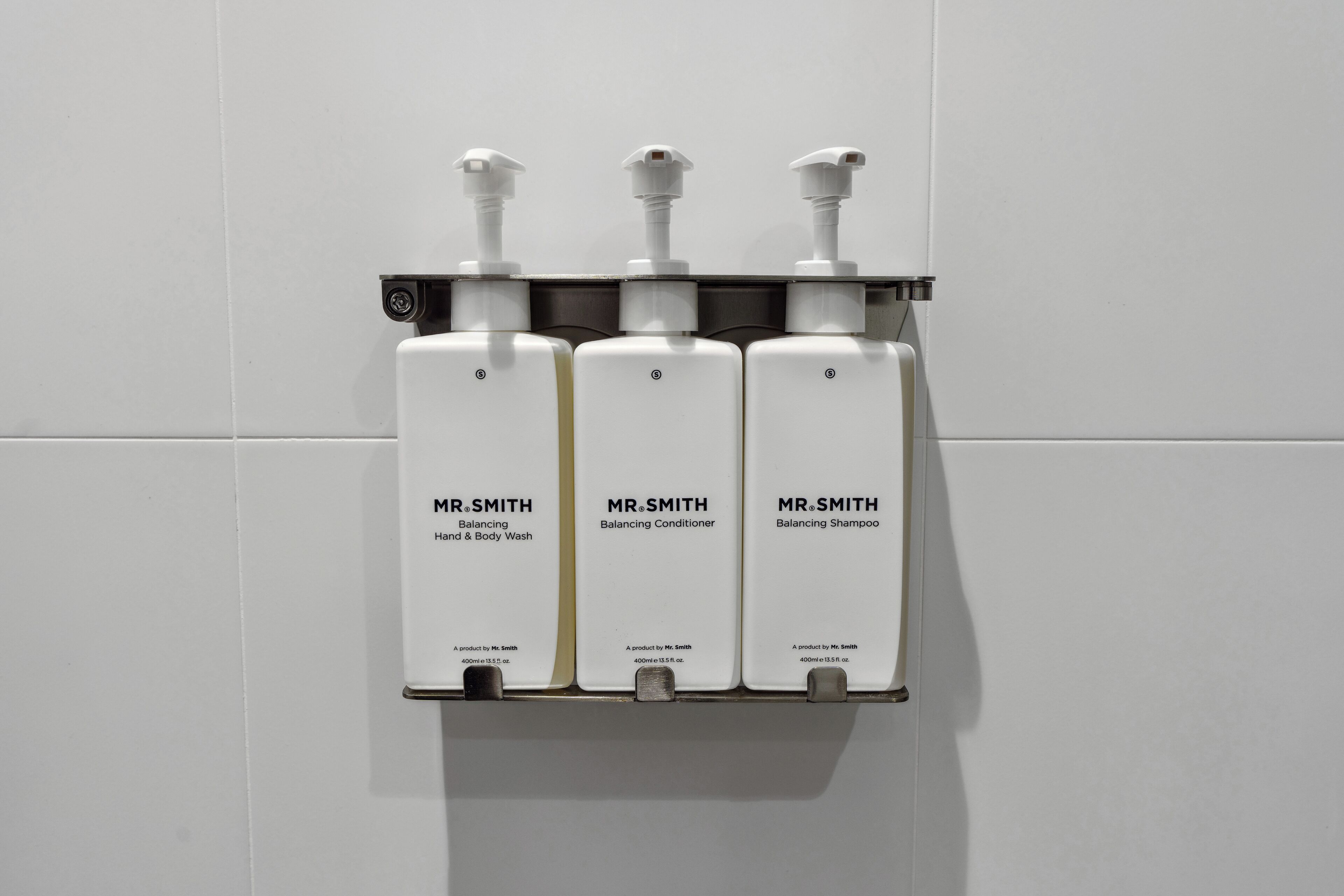 A wall-mounted dispenser with three bottles labeled Mr. Smith products: hand and body wash, conditioner, and shampoo.