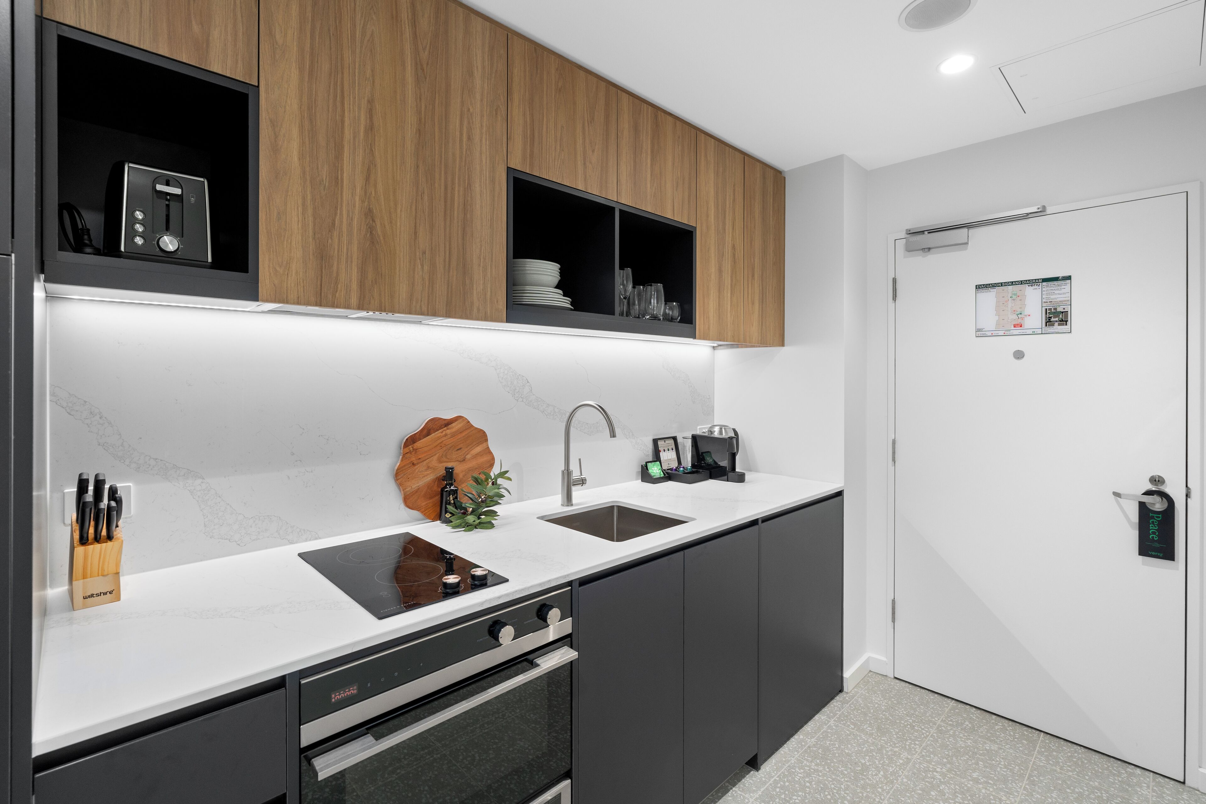 A modern kitchen with a built-in oven, a sink, and a microwave. There are wooden cabinets above the counter and a door with a lock on the right.