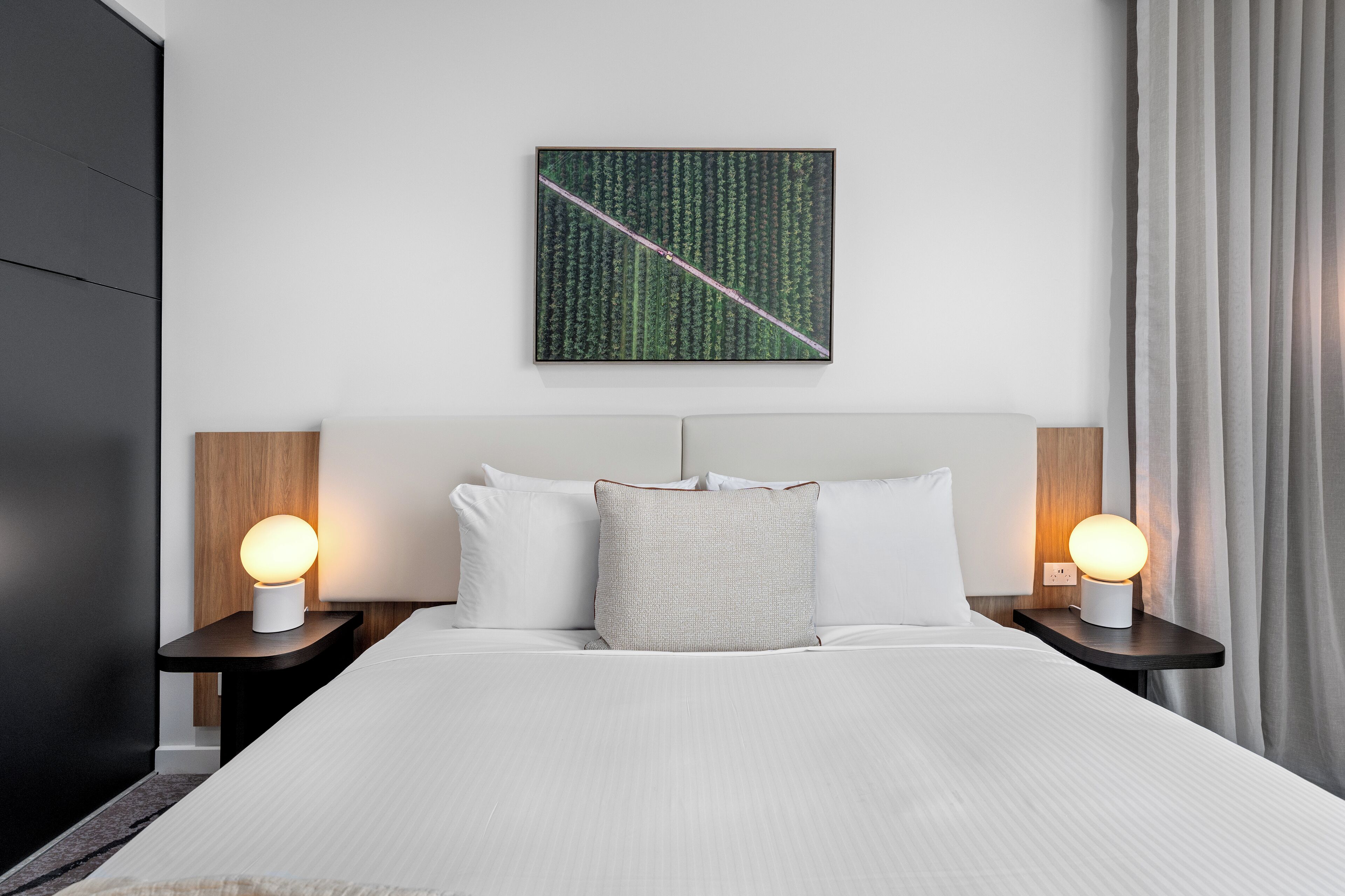 A modern hotel room with a neatly made bed, two bedside tables with lamps, a large abstract painting on the wall, and a view of the outdoors through the window.
