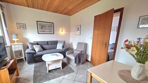 A living room with a sofa, armchair, coffee table, TV, and a dining area with a table and chairs. - Haus Dorfschmiede, Whg. Seehund - Haus Dorfschmiede (Alkersum)