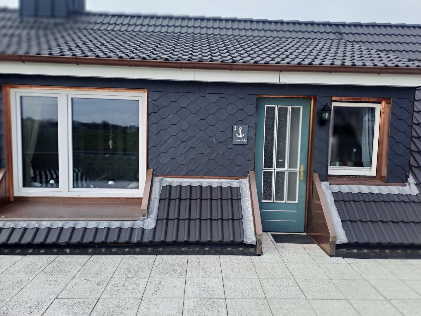 Un bâtiment avec un toit recouvert de tuiles sombres, une porte verte et une fenêtre dotée d’un cadre blanc.