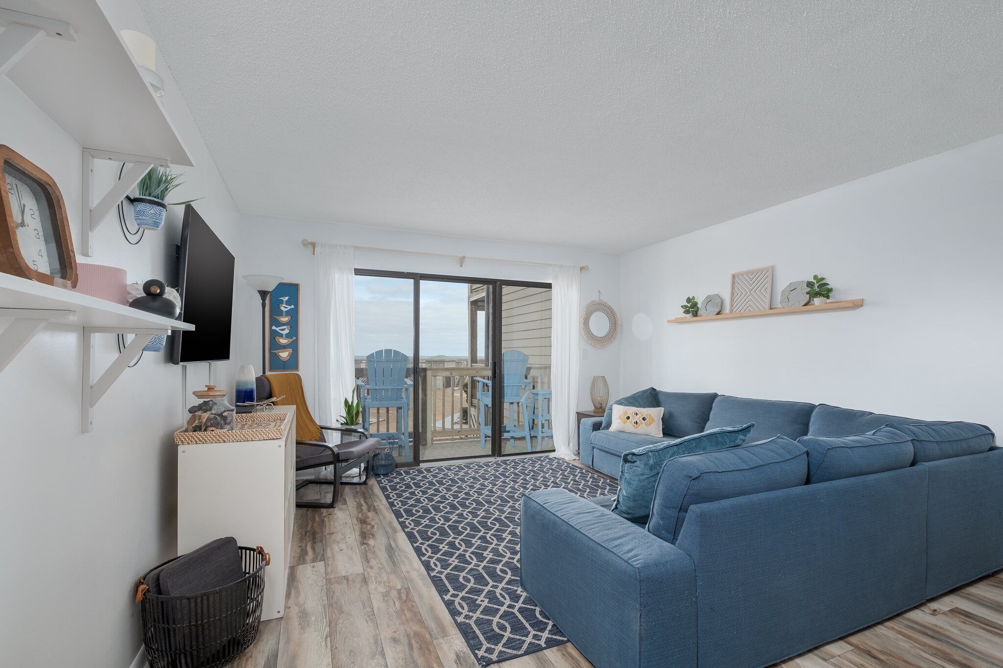 A modern living room with a blue sofa, a flat-screen TV, and a large sliding glass door leading to a balcony.