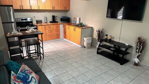 A compact kitchen with wooden cabinets, a dining area with a table and chairs, a black TV on the wall, and a tiled floor.