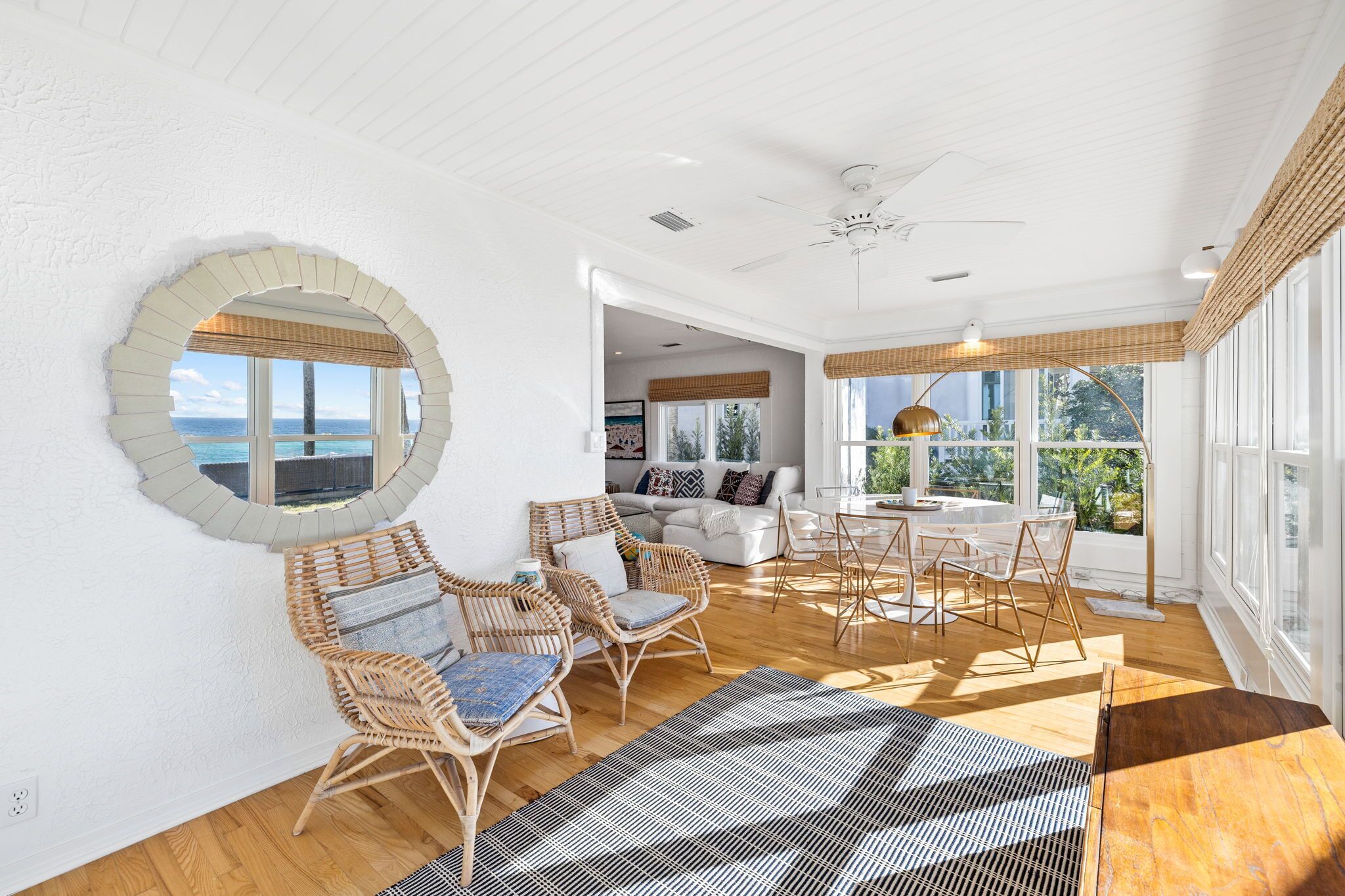 A spacious room with a large window offering a view of the sea, wicker furniture, a ceiling fan, and a dining area with a table and chairs.