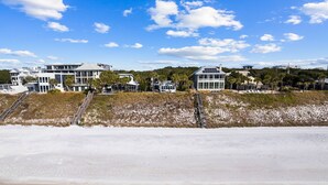 Een pand aan het strand met meerdere gebouwen, waaronder een groot huis met een glazen gevel en een kleinere constructie. Er zijn trappen die naar het strand leiden, en het gebied is omringd door begroeiing.