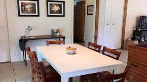 Een eethoek met een witte tafel en houten stoelen, een plafondventilator en lijstjes met afbeeldingen aan de muur.