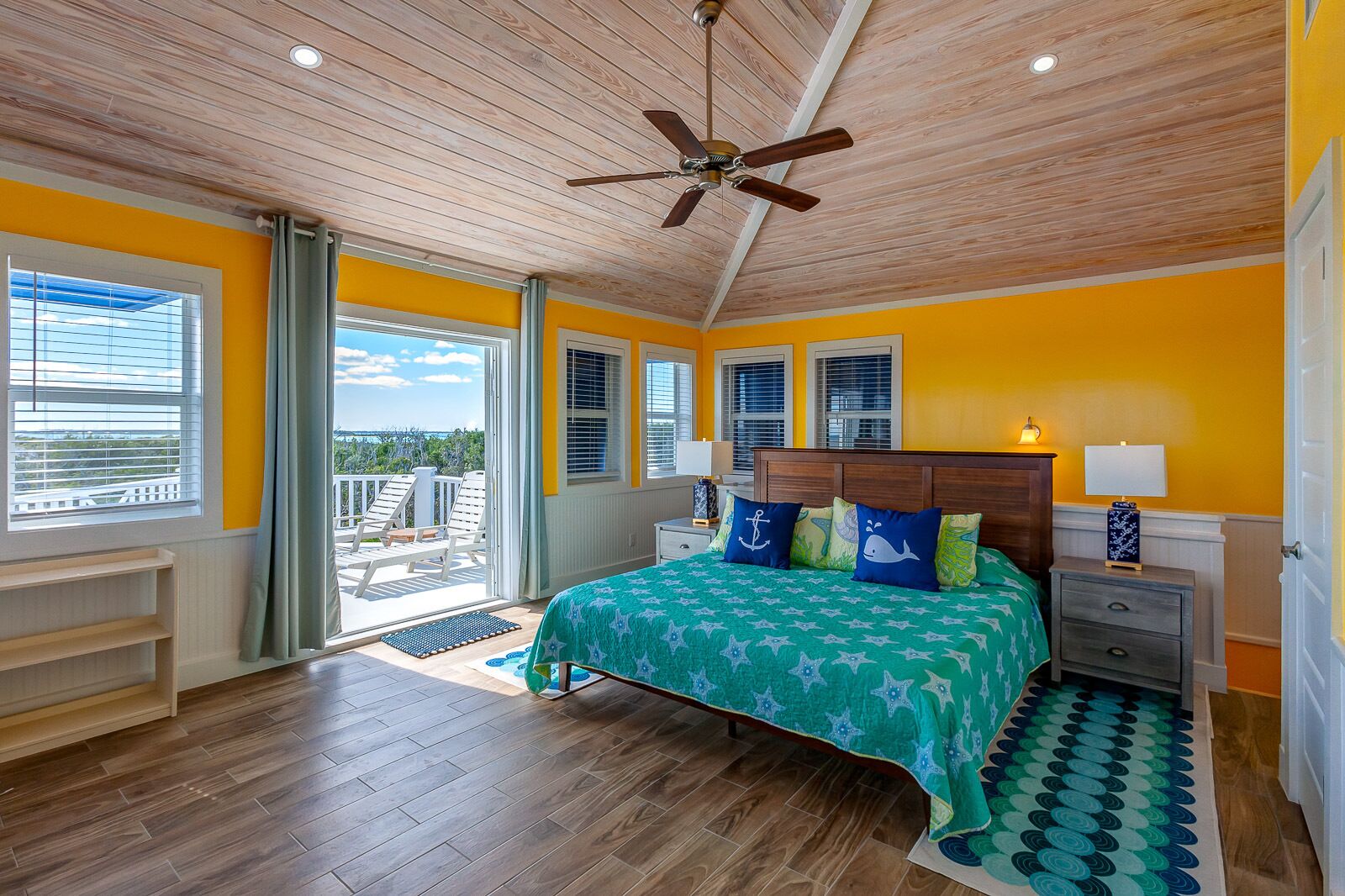 A bedroom with a wooden ceiling, a bed with a green and blue bedspread, a nightstand with a lamp, and a view of the outdoors through a sliding door.