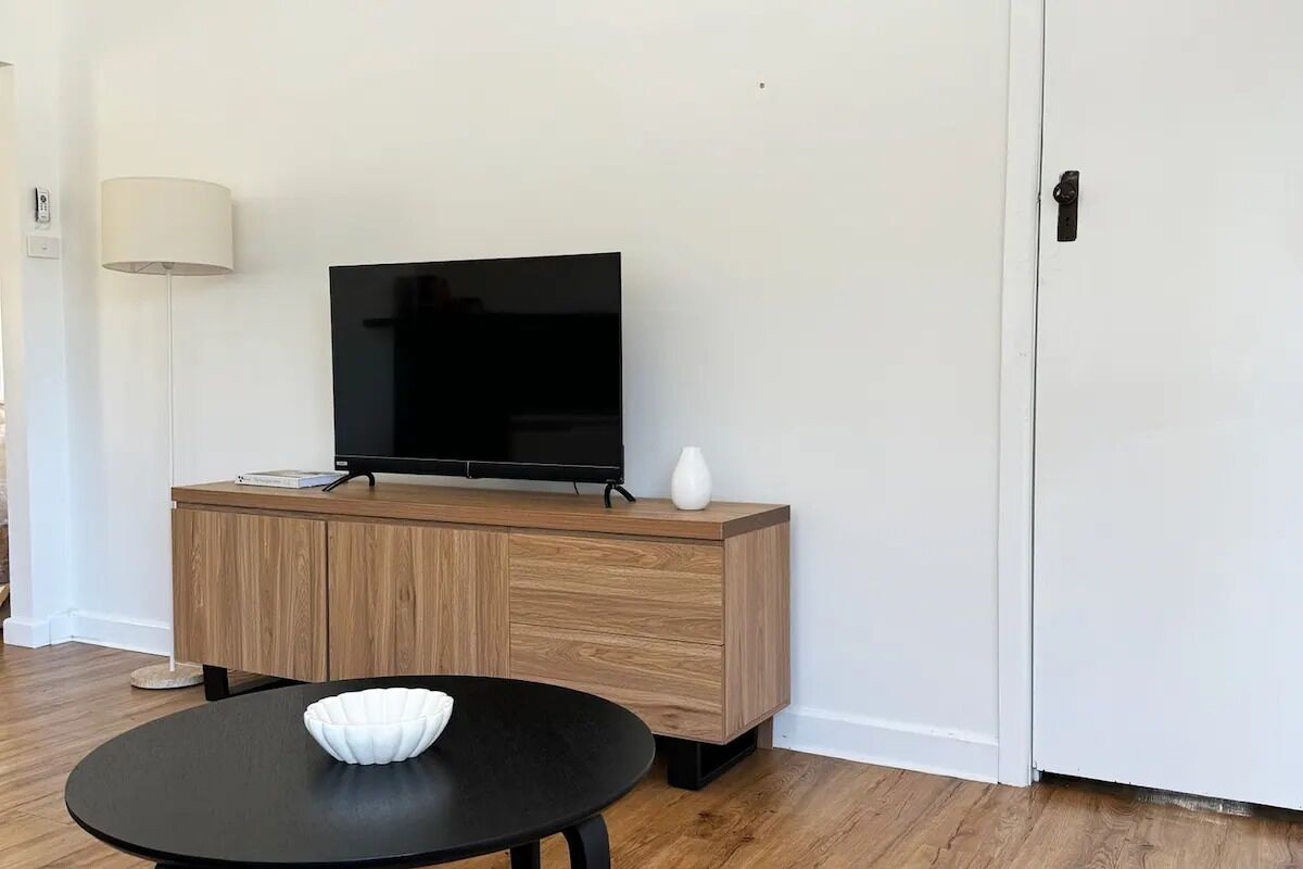 Una sala de estar moderna con una televisión de pantalla plana sobre un mueble de madera, una mesa redonda negra con un cuenco blanco y una lámpara de pie.