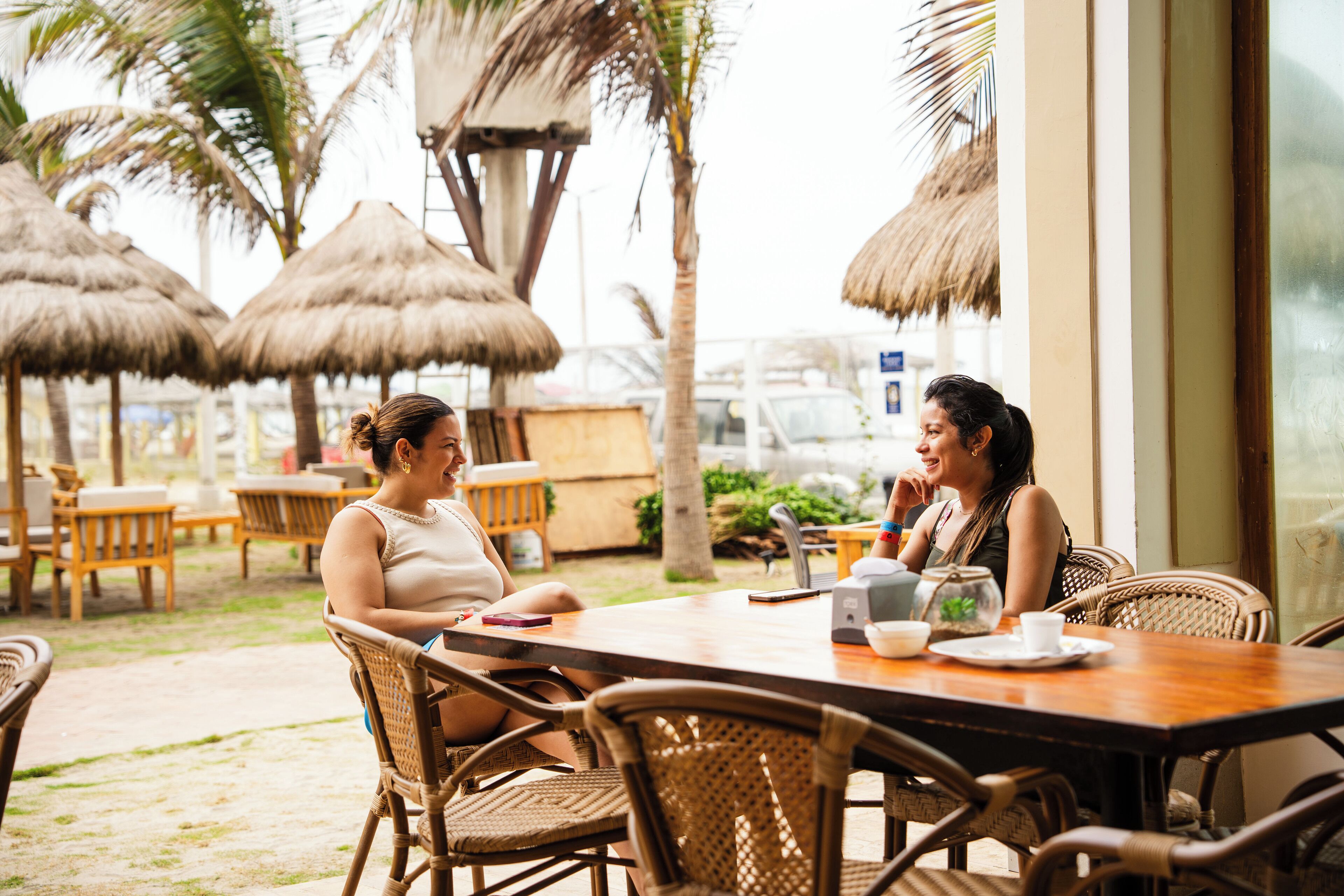 Two people seated at a wooden table with a view of a beach and palm trees in the background.