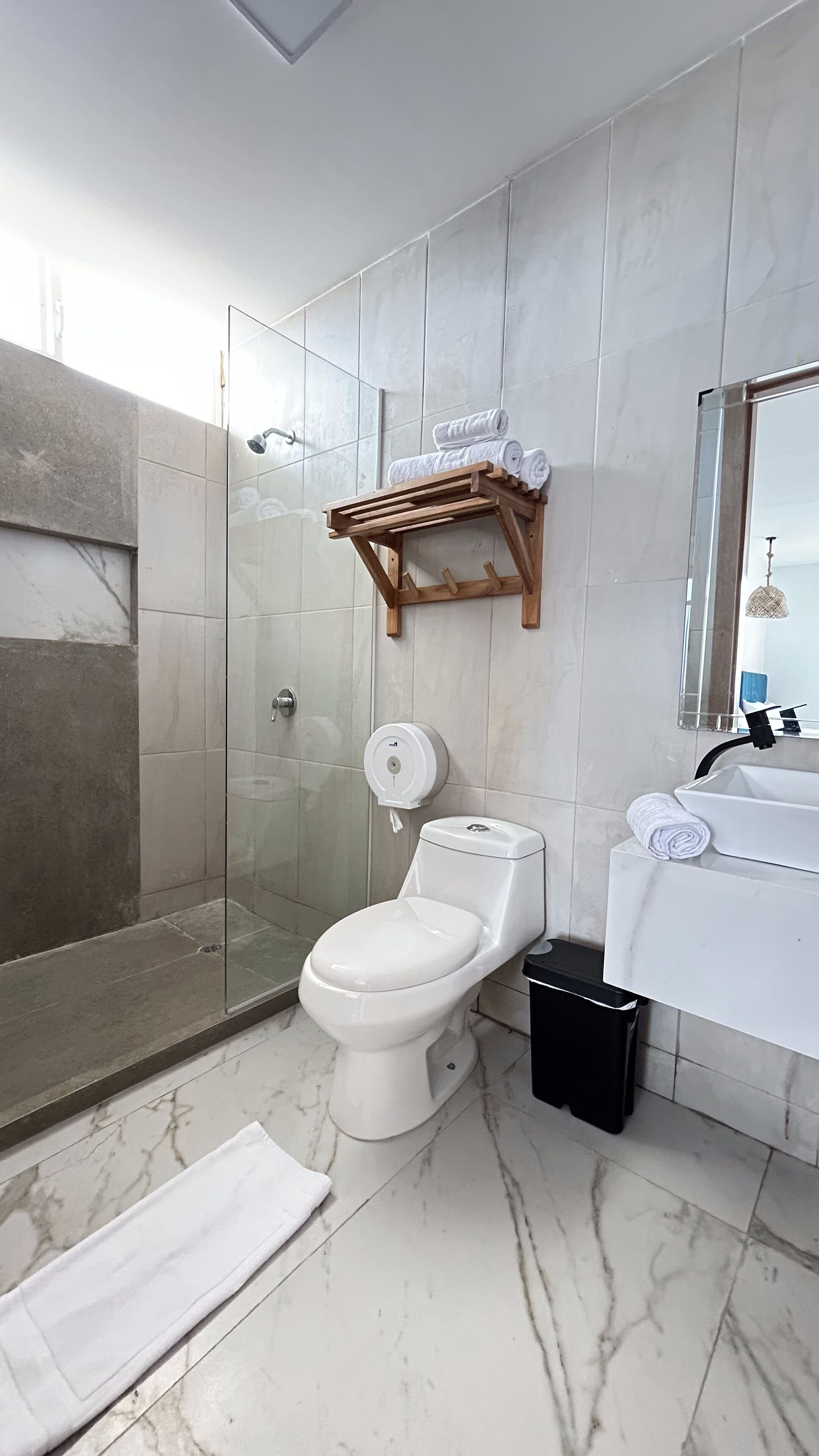 A bathroom with a toilet, a hand dryer, a glass shower enclosure, a wooden towel rack, and a black trash bin.