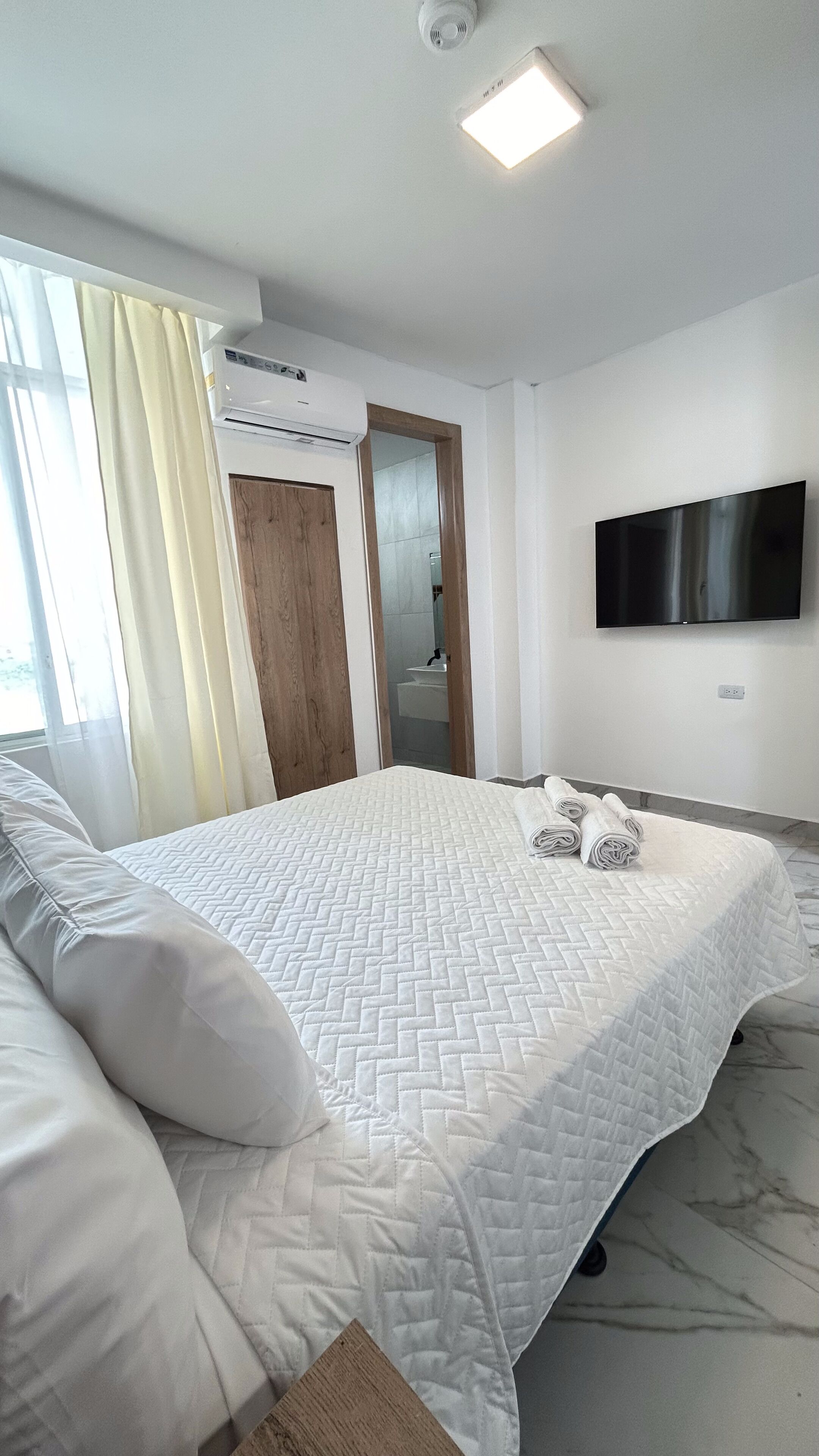 A modern hotel room with a large bed, a flat-screen TV mounted on the wall, and an air conditioning unit on the ceiling.