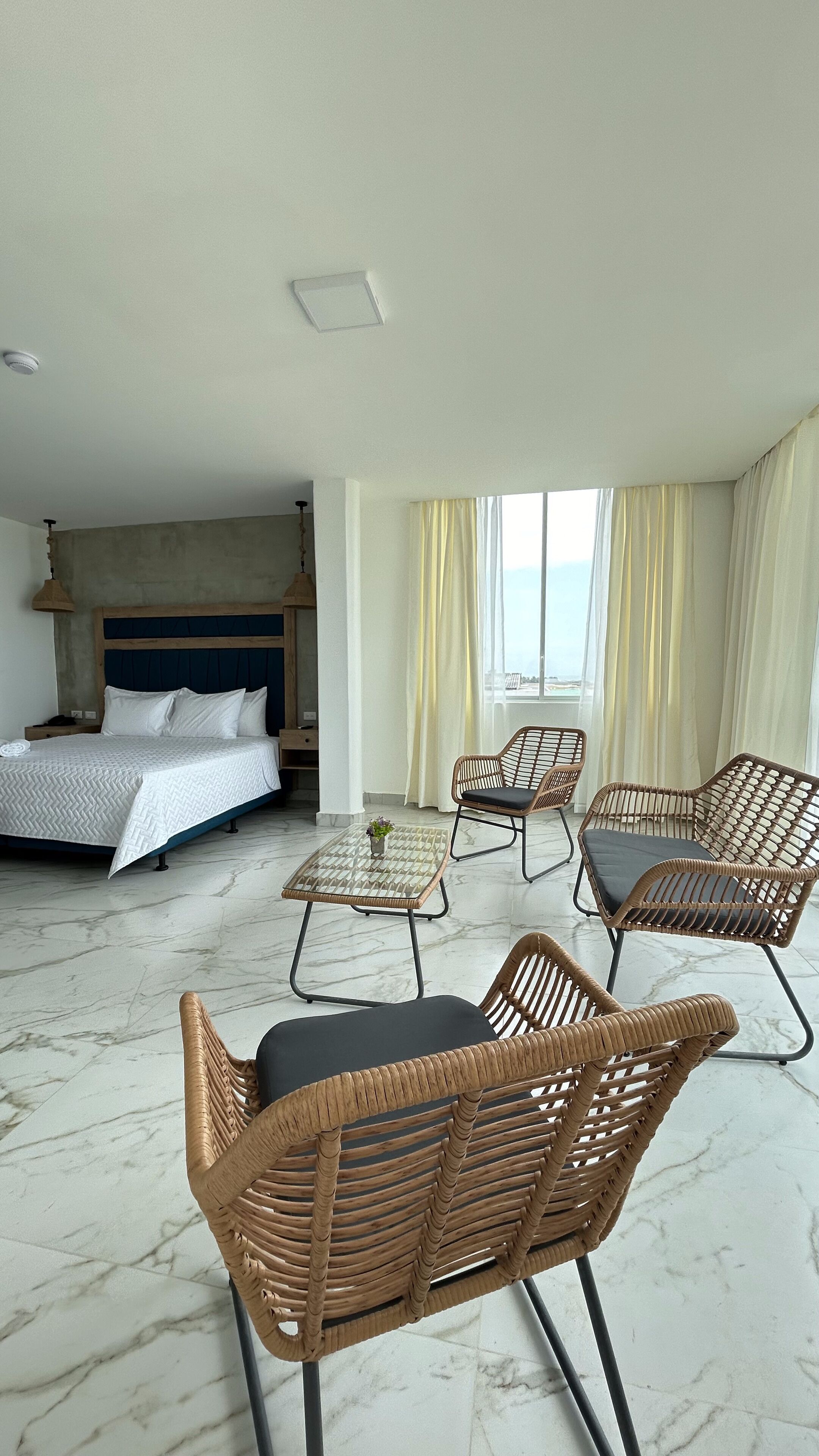 A hotel room with a bed, two wicker chairs, a small table, and a view of the sea through large windows.