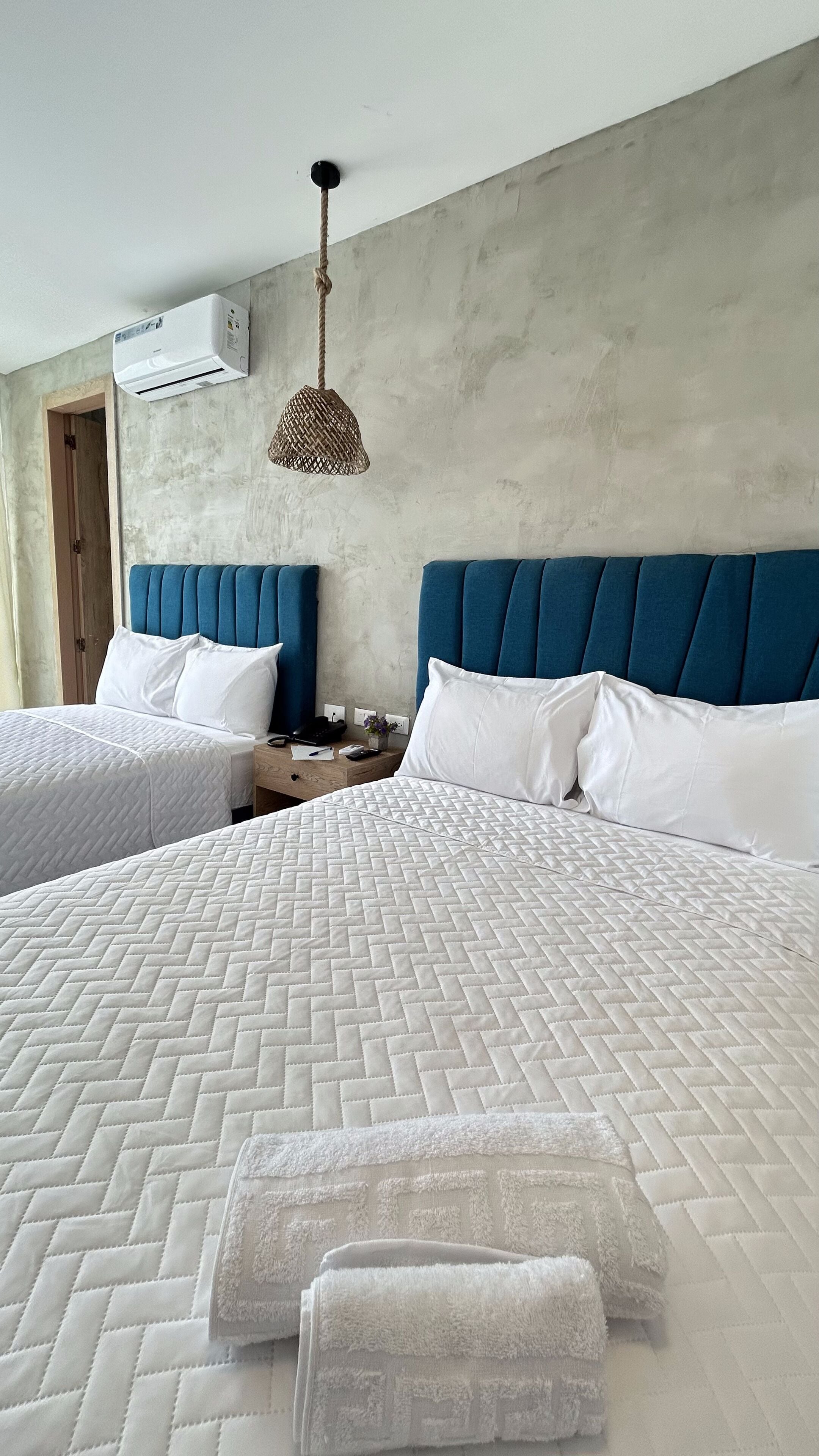 Habitación de hotel con dos camas, cada una con colchas blancas y cabeceros azules. Se observa un lámpara colgante y un equipo de aire acondicionado en la pared.