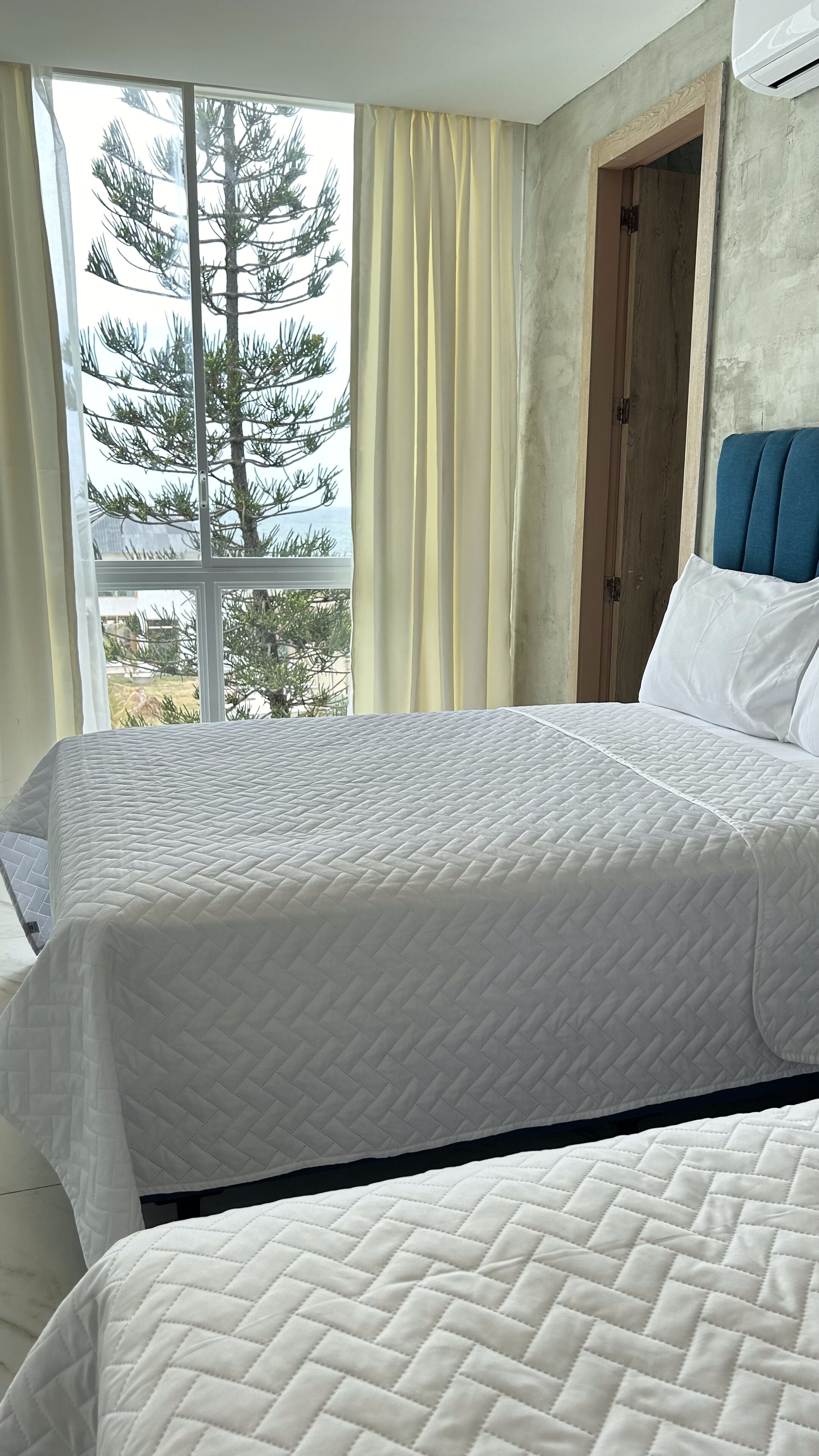 A hotel room with two beds, a large window with curtains, a view of trees, and a wooden door.