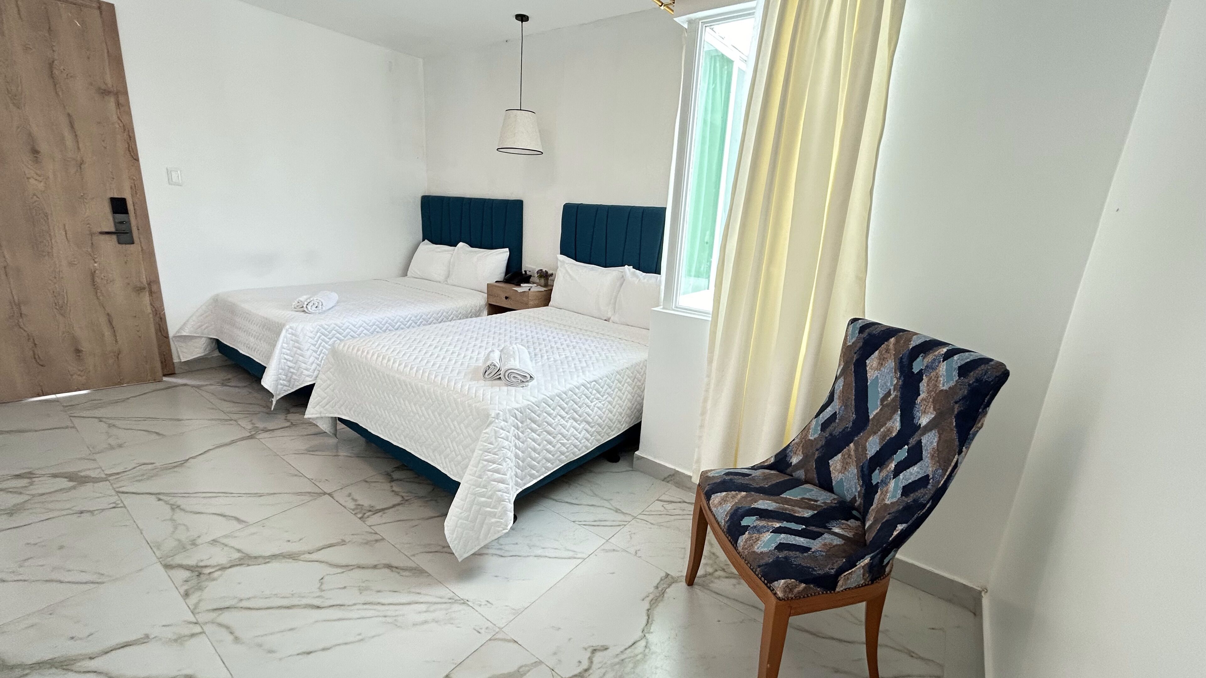 A room with two beds, a chair, a wooden door, and a window with curtains.