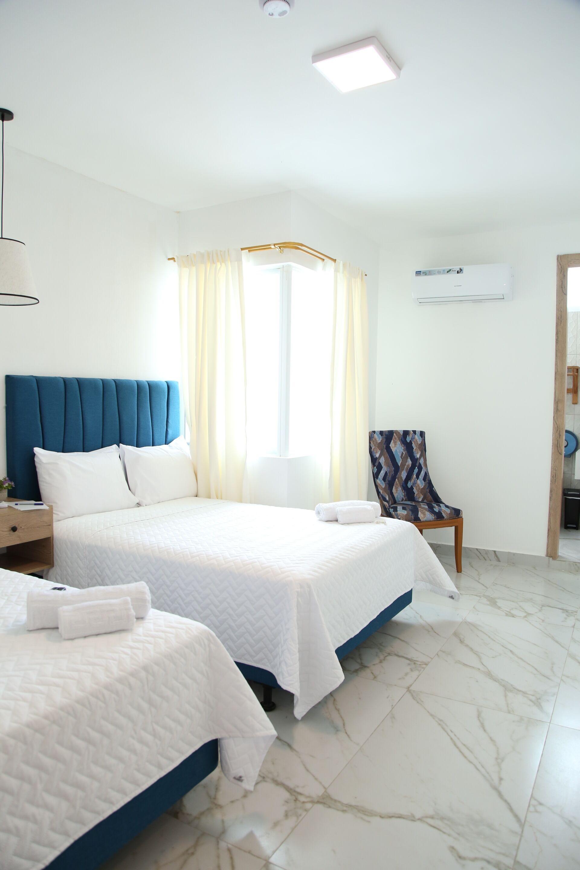 Habitación de hotel con dos camas, una silla, una mesita de noche y un ventanal con cortinas.