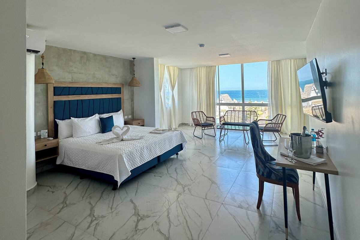 Ein Hotelzimmer mit Bett, Esstisch mit Stühlen, Fernseher und Blick auf den Strand.