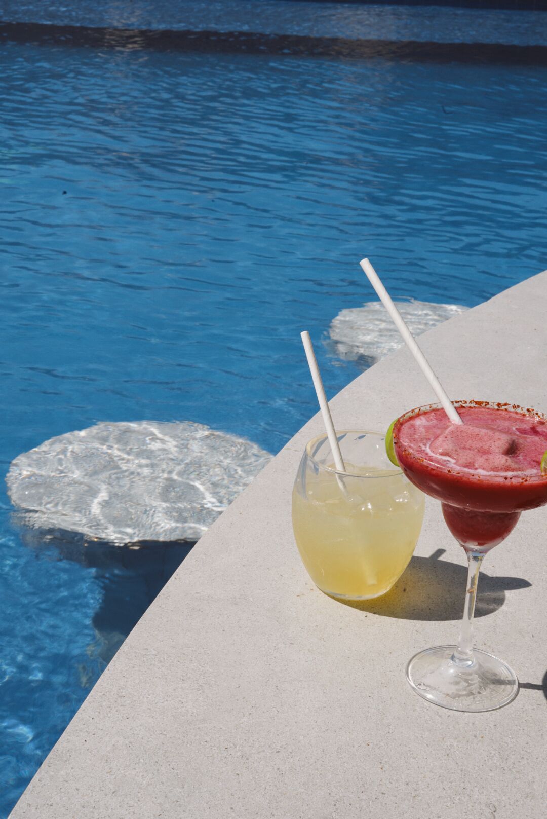 A poolside area with a concrete edge, two drinks in glasses, and a partially submerged ice block.