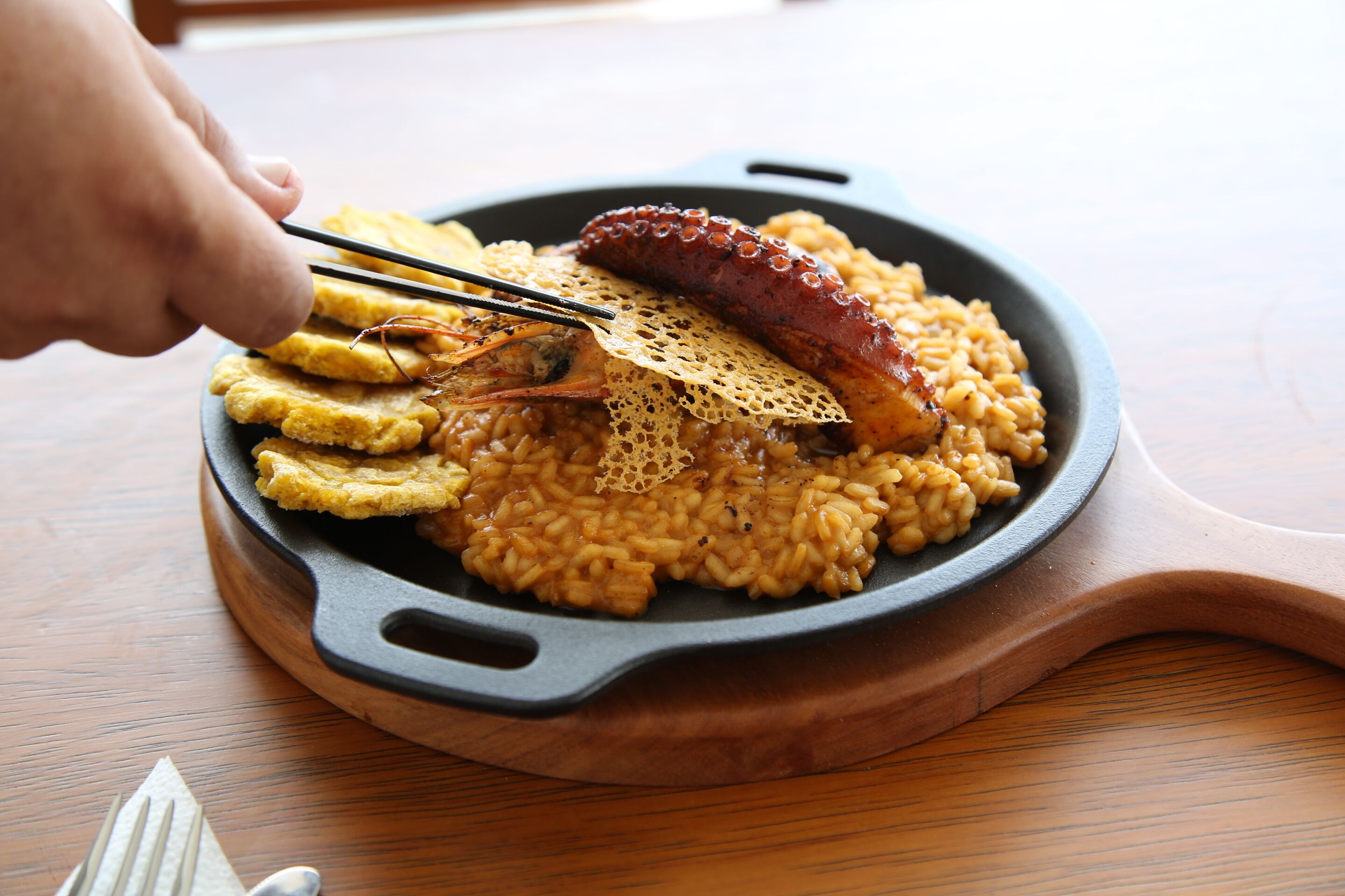 A skillet with a sausage and rice, a wooden serving board, and a hand using chopsticks.