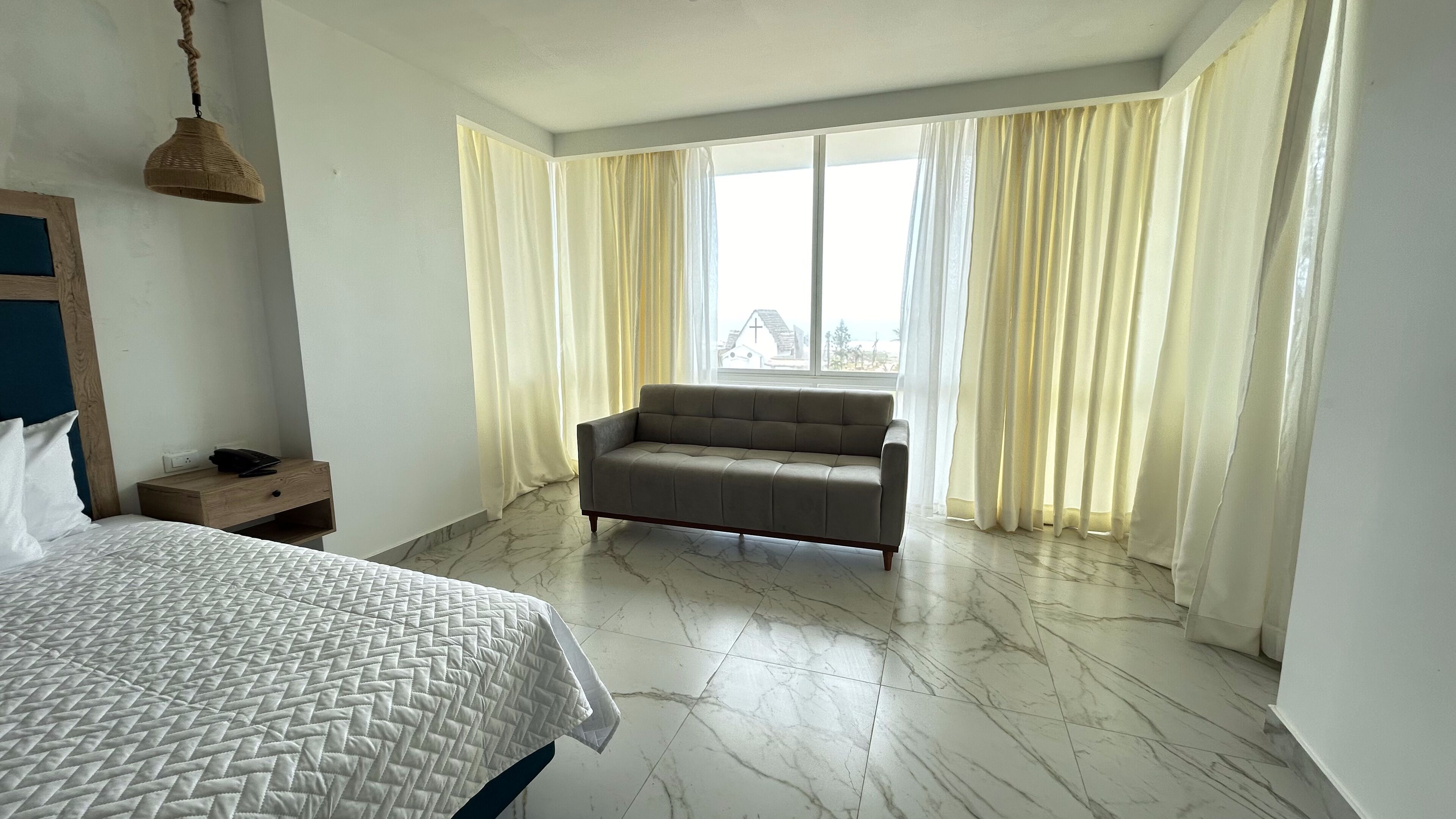 A hotel room with a bed, a sofa, a wooden nightstand, and a large window with curtains.