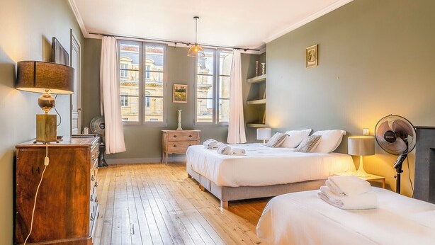 Une chambre avec deux lits, une commode en bois, une lampe, un ventilateur et une fenêtre donnant sur des immeubles.