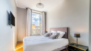 Une chambre à coucher avec un lit, une table de chevet, une lampe, un téléviseur fixé au mur et une fenêtre avec des rideaux.