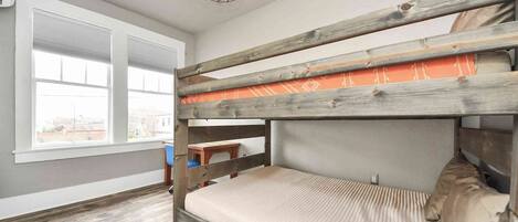 A bunk bed with a wooden frame and a patterned mattress, a small wooden table, and a window with a view of buildings.