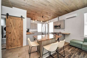 A modern kitchen with a central island, white bar stools, a wooden barn door, and a green sofa.