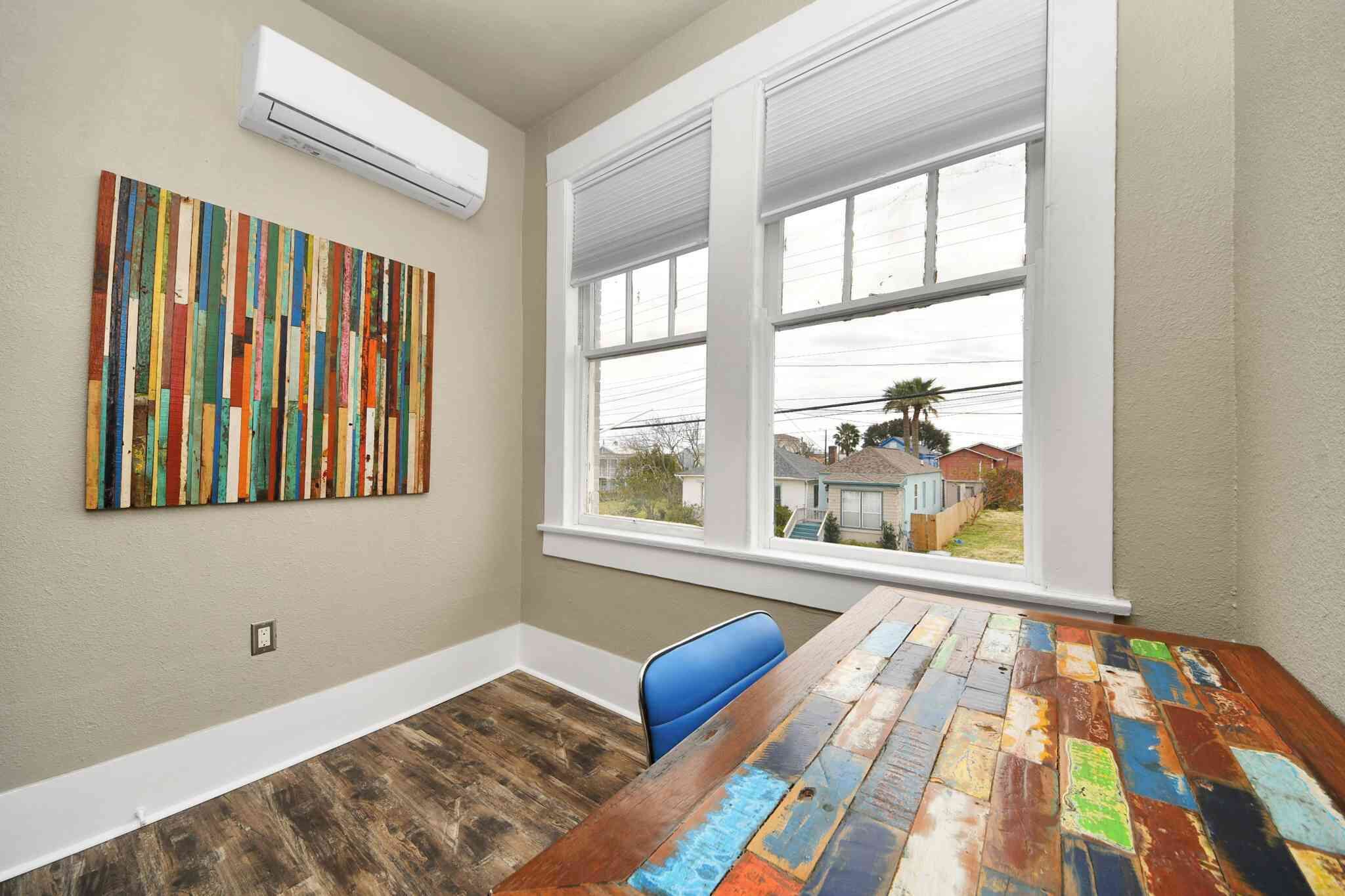 A room with a wooden table, a blue chair, a colorful abstract painting, and a window with a view of houses.