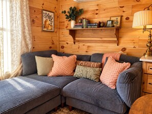 Ein gemütliches Blockhauszimmer mit einer Eckcouch, einem Holzregal mit Büchern und Dekoration sowie einer Lampe.