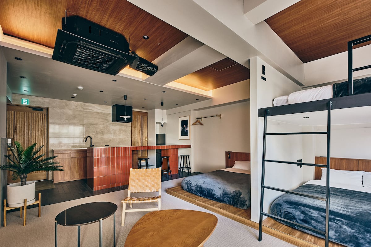 A modern hotel room with a bunk bed, a small kitchen area, a seating area with a table and chairs, and a potted plant.