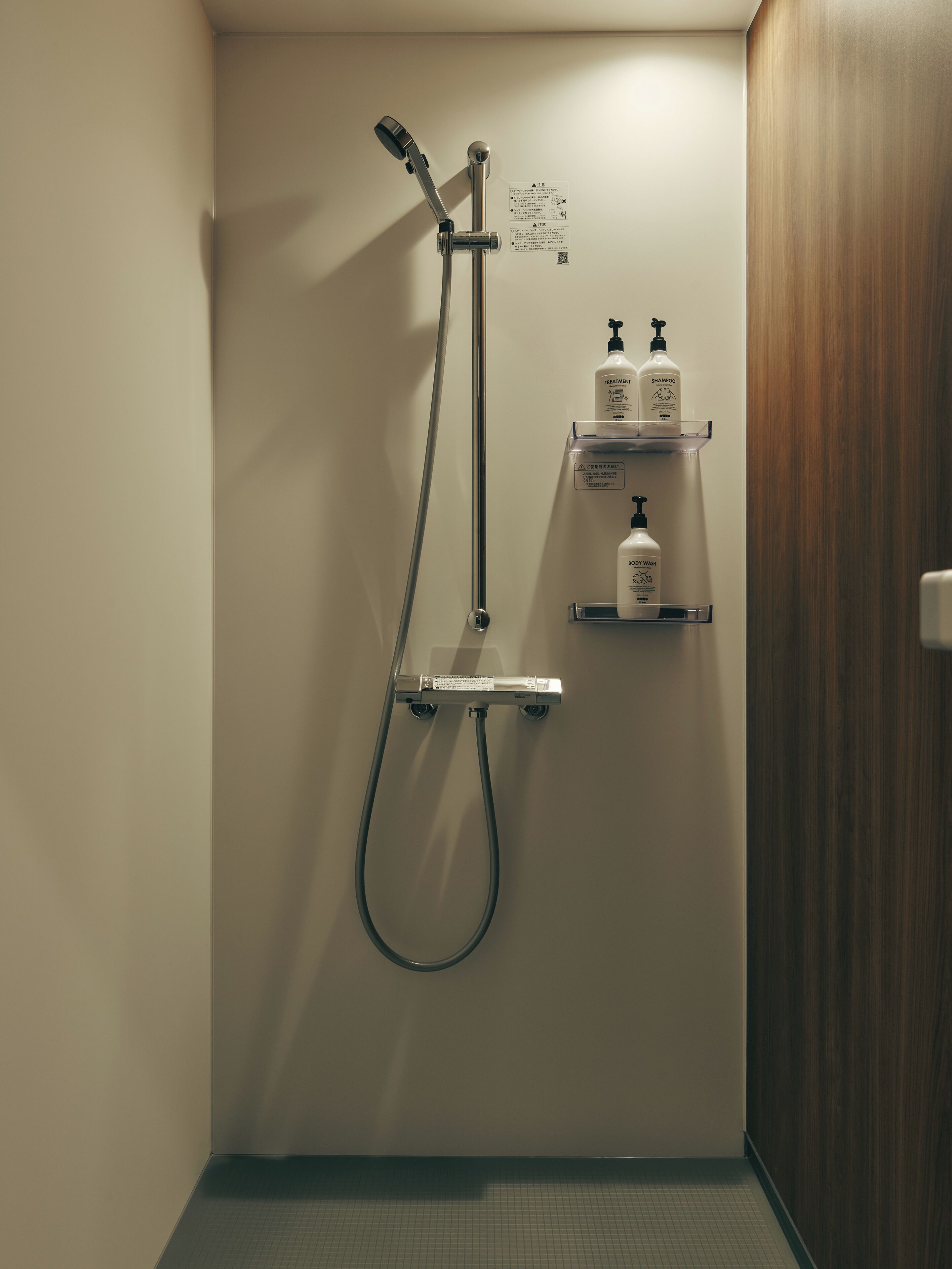 A shower with a handheld showerhead and a wall-mounted faucet, a small shelf with toiletries, and a wooden door.