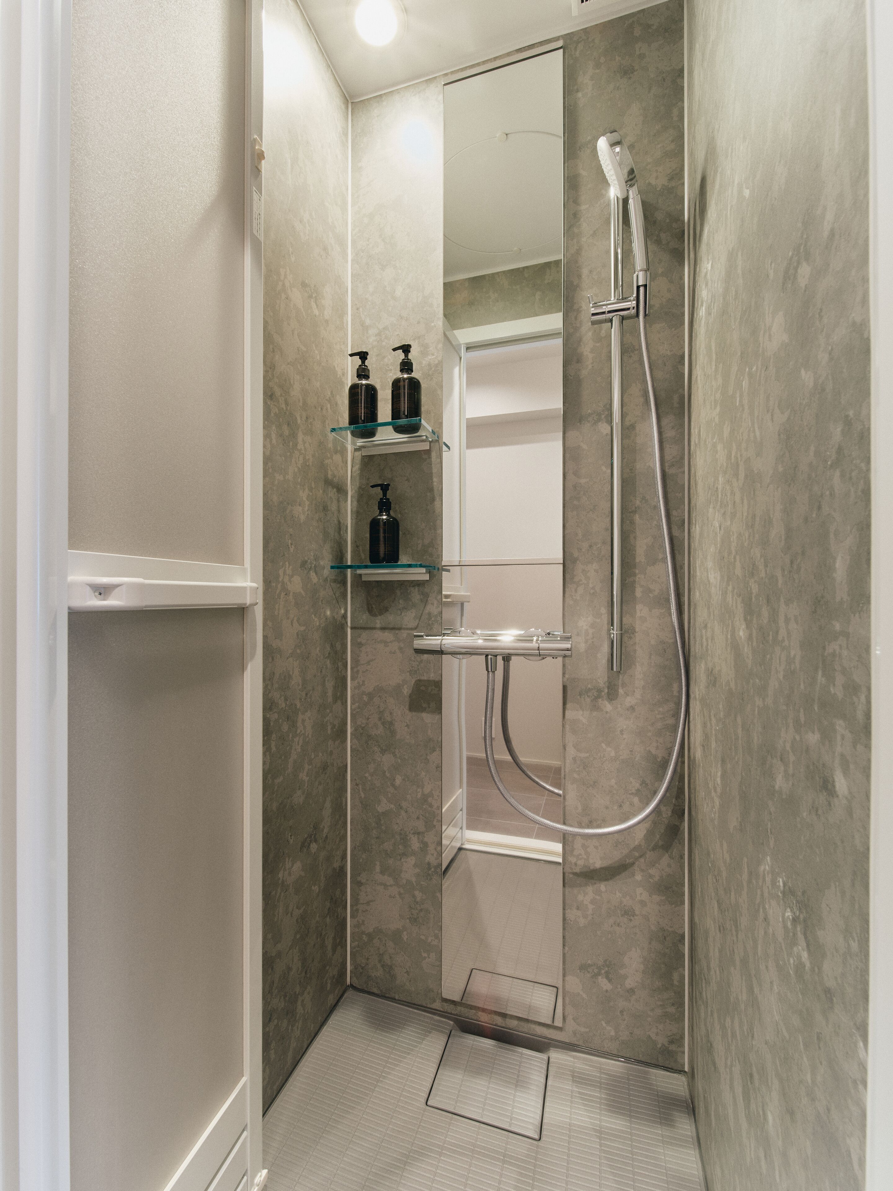 A modern shower stall with a glass door, a handheld showerhead, and a handheld shower attachment. There are two shelves with bottles, likely containing shampoo and conditioner.