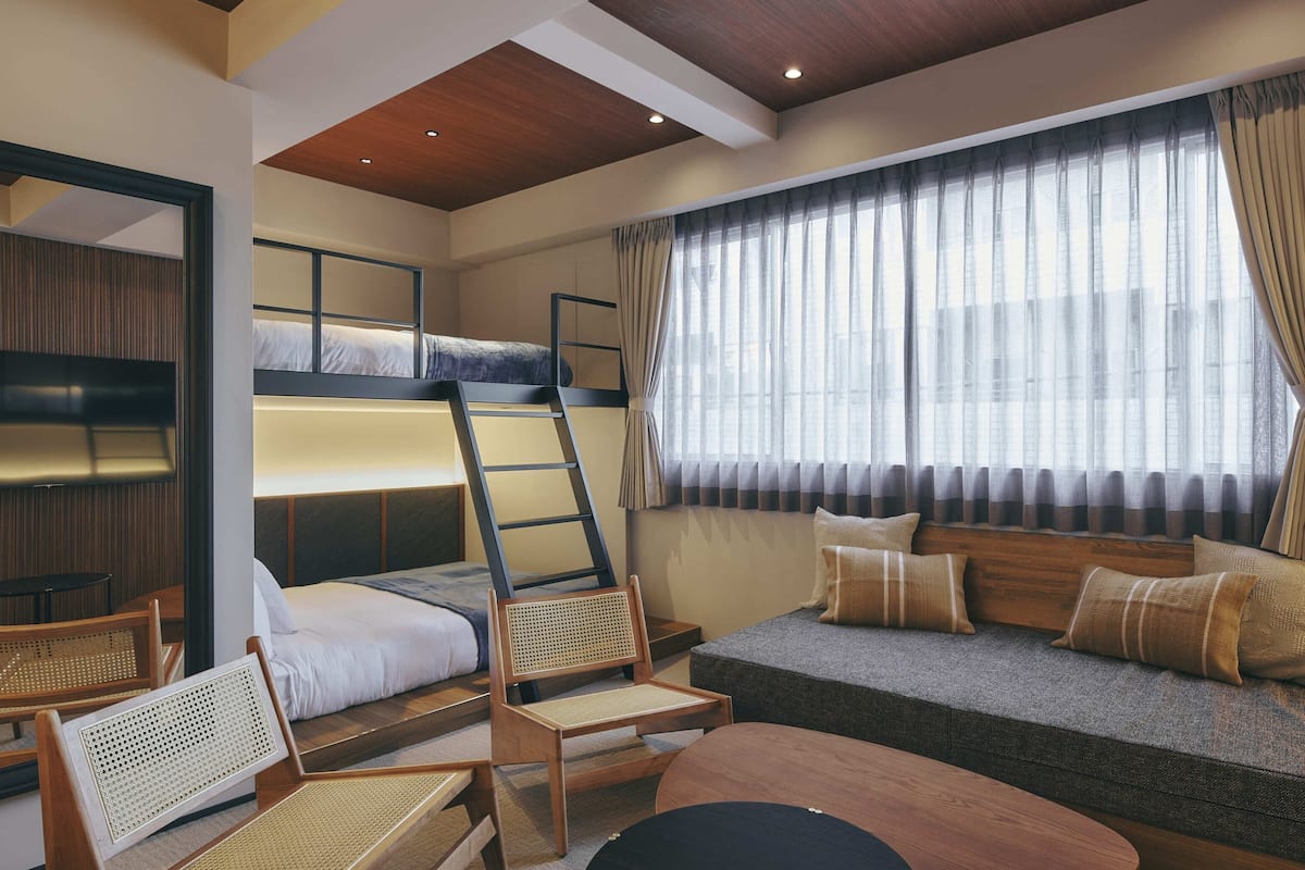 A bunk bed room with a ladder, a sofa, two chairs, and a window with curtains.