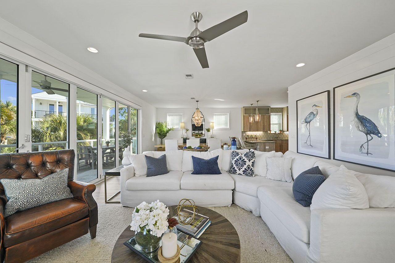 A spacious living room with a white sectional sofa, a leather armchair, and a coffee table. There is a ceiling fan, large windows, and artwork on the walls.