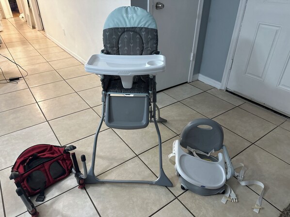 Un cochecito de bebé con una mesita, una mochila roja y una sillita gris sobre un suelo de baldosas.