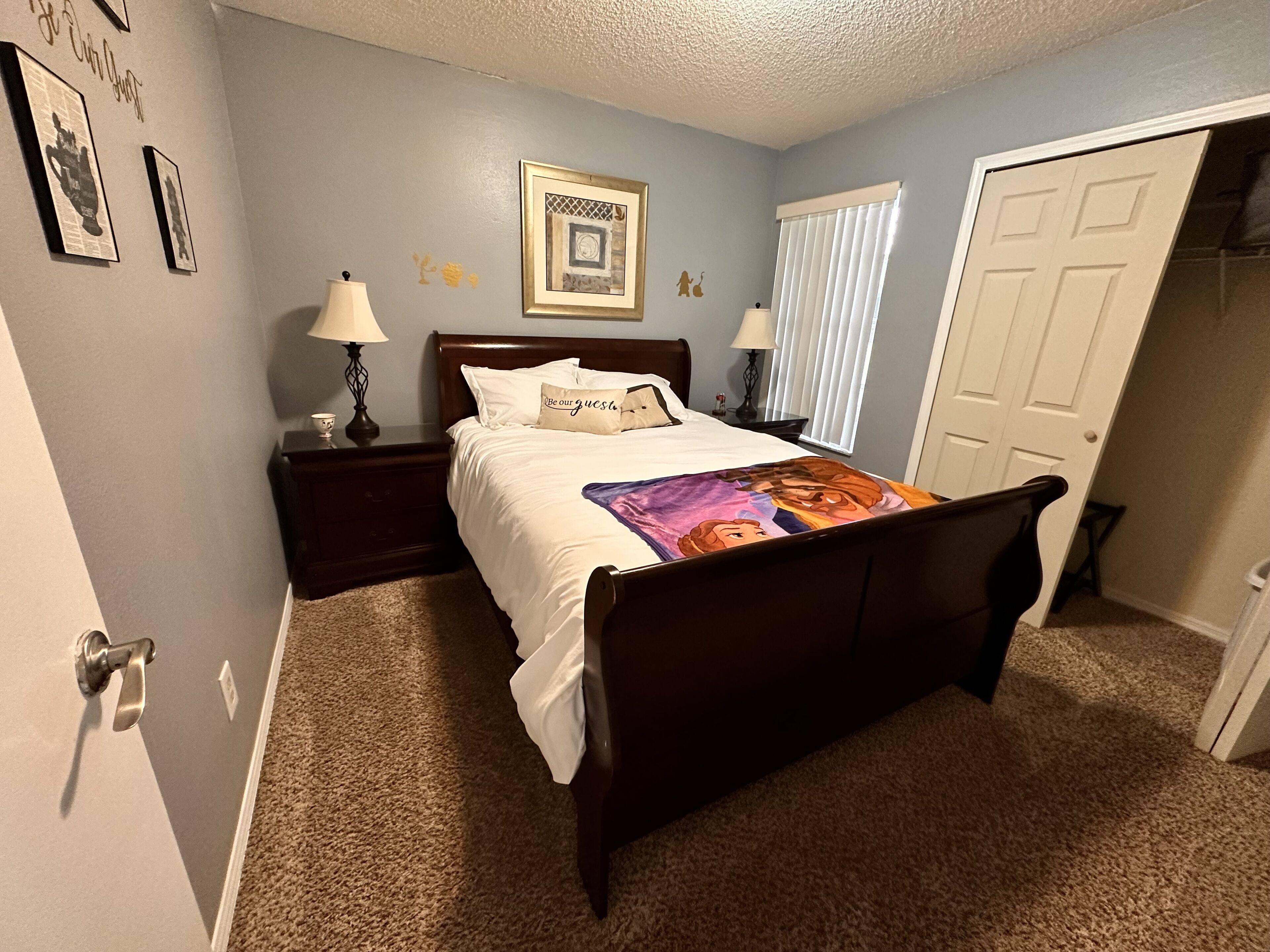 A bedroom with a bed, nightstands, a lamp, a door, and a wall with framed pictures.