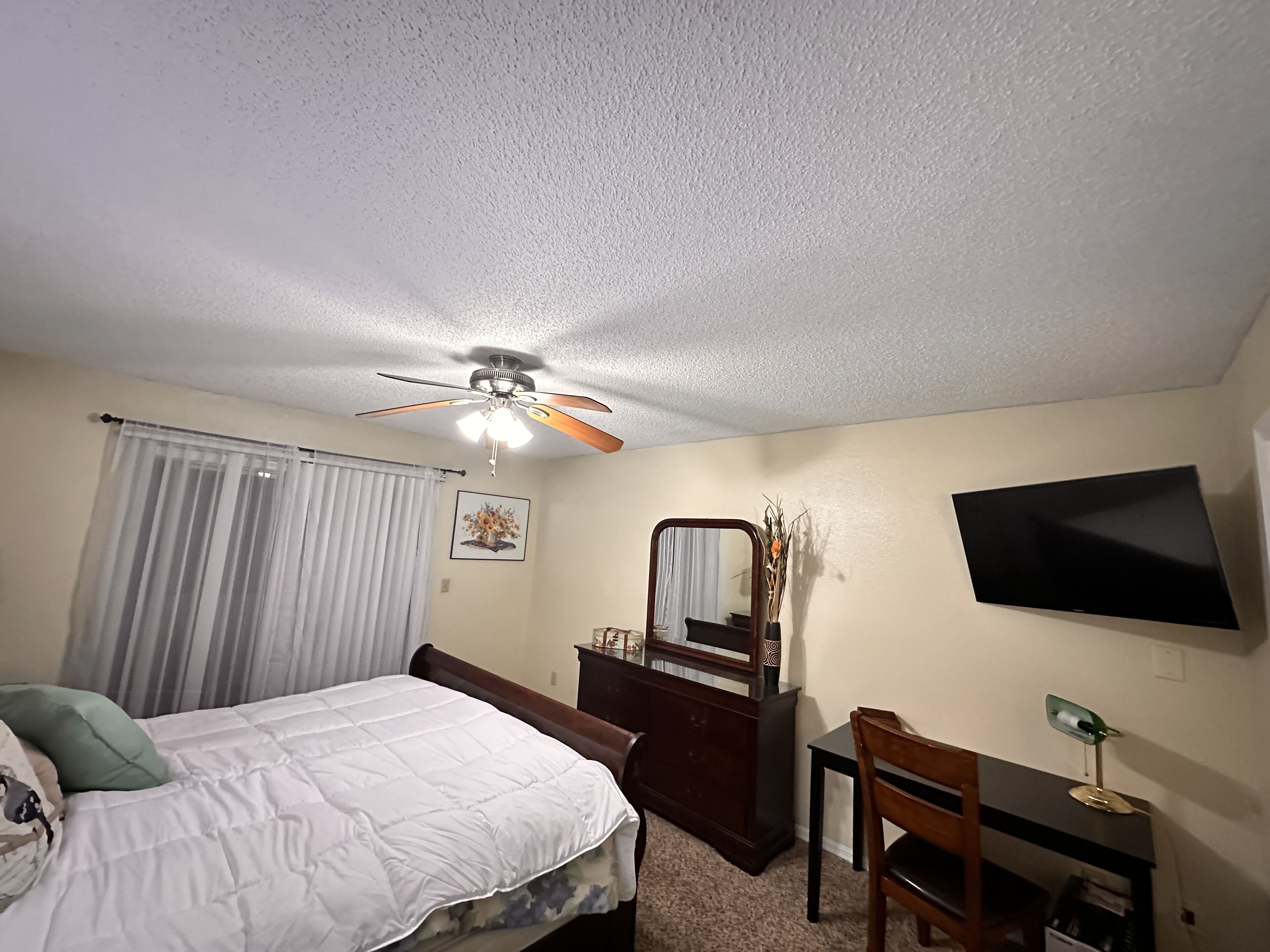 A bedroom with a bed, a desk, a chair, a dresser, a mirror, a ceiling fan, a TV, and a window with curtains.