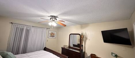 A bedroom with a bed, a desk, a chair, a dresser, a mirror, a ceiling fan, a TV, and a window with curtains.
