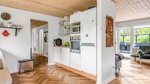 Une cuisine moderne avec des armoires blanches, un four encastré et un four à micro-ondes. Le sol est en bois, disposé en motif de parquet, et le plafond est également en bois. On peut voir une horloge au mur.