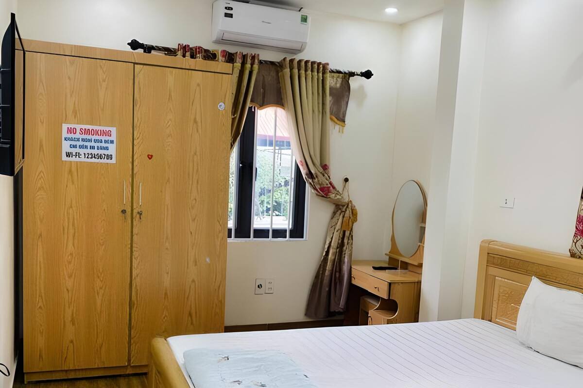 A hotel room with a wooden wardrobe, a bed with a white sheet, a bedside table with a mirror, and a window with curtains.