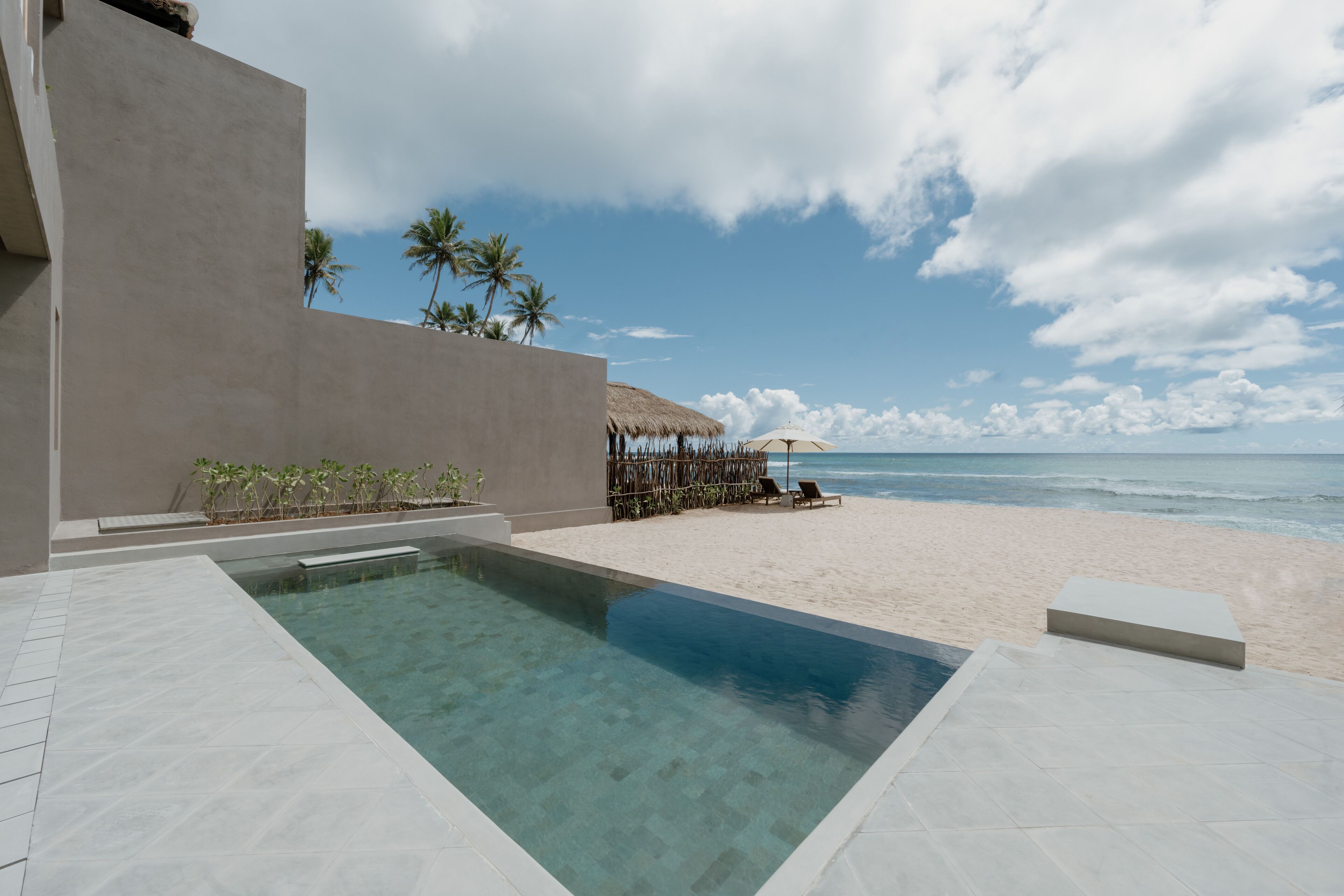 A swimming pool adjacent to a beach with a thatched-roof structure and lounge chairs.