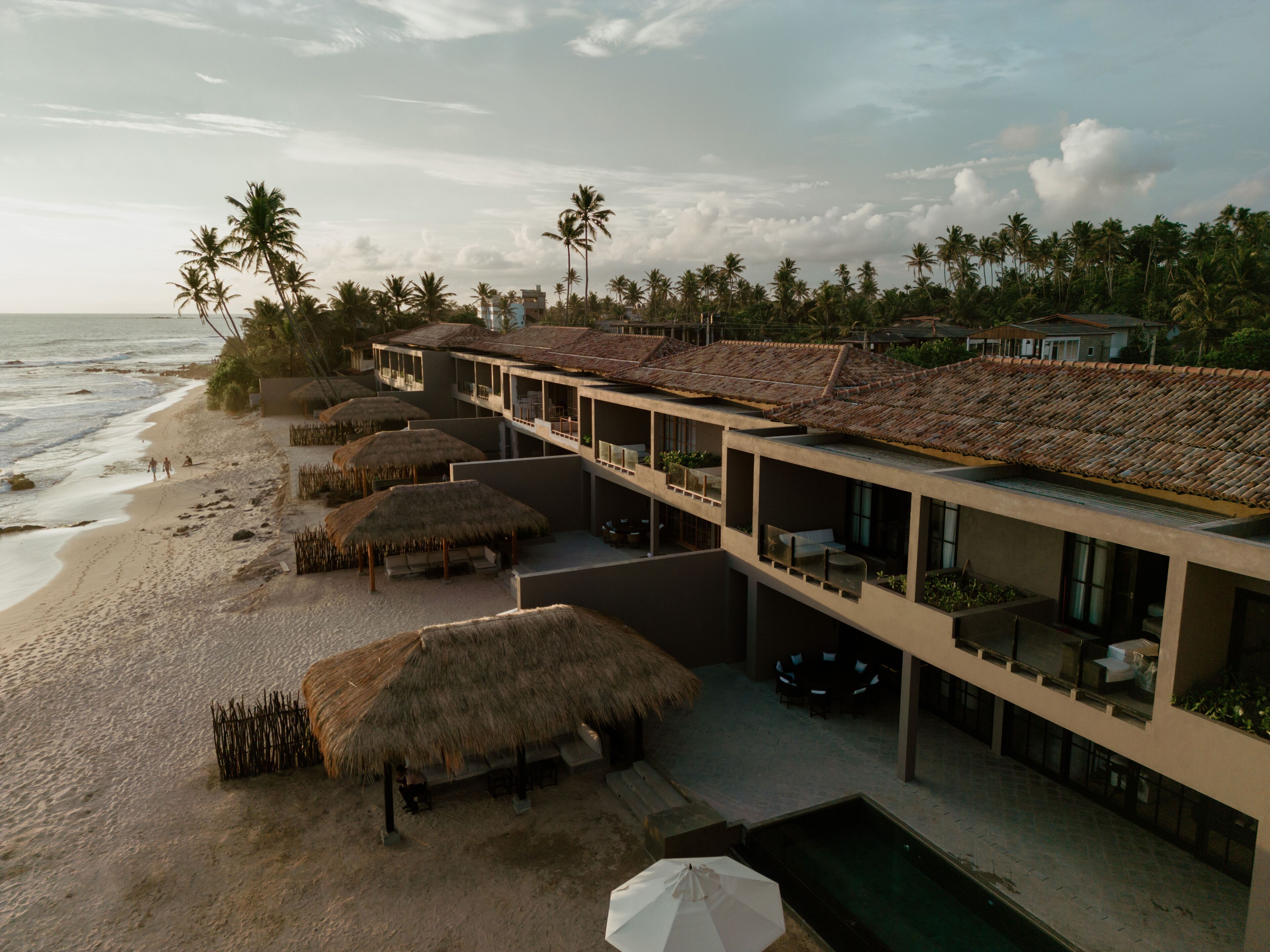 A beachfront hotel with multiple balconies overlooking the ocean, thatched umbrellas, and a swimming pool.
