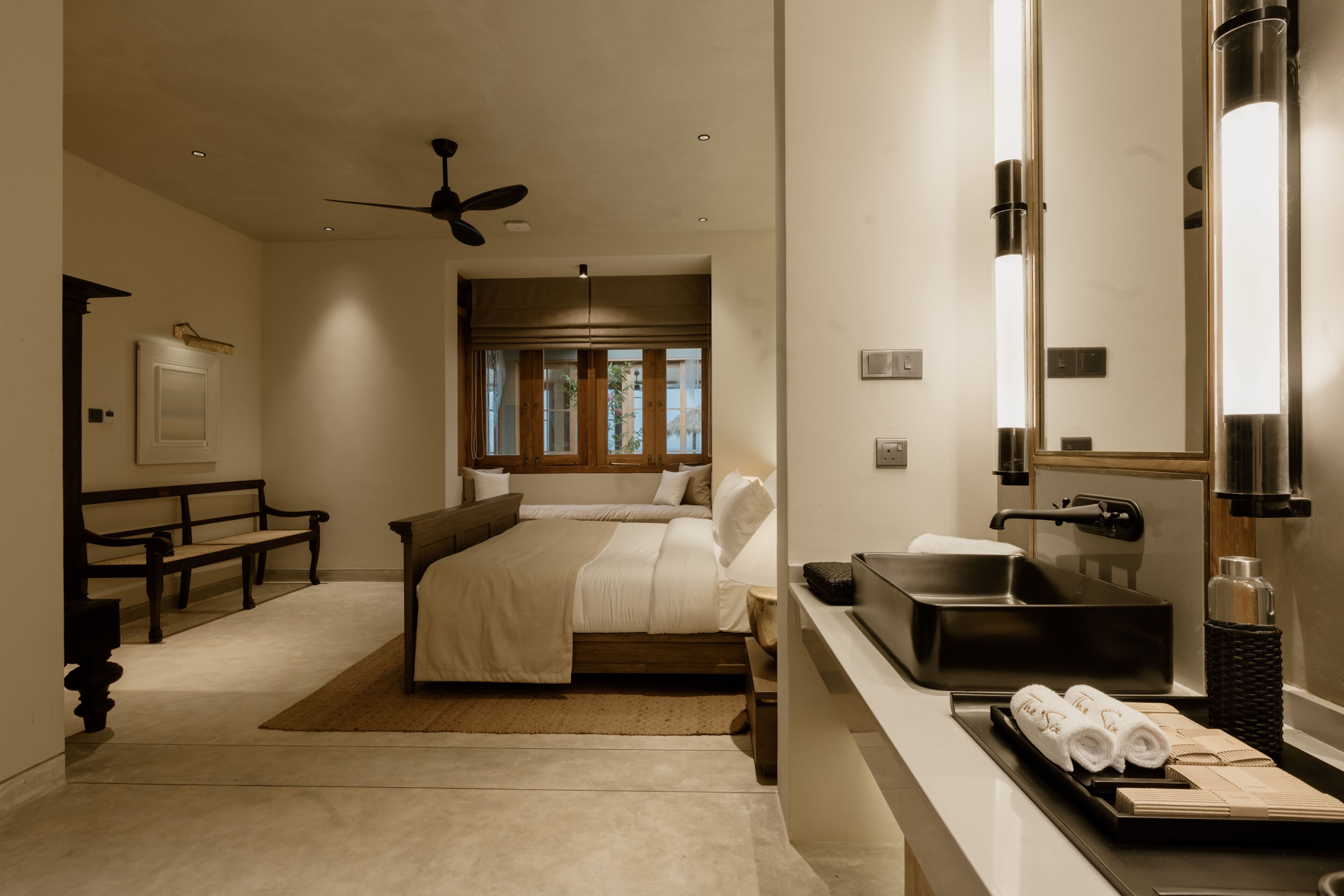A modern bedroom with a bed, a bench, a desk, and a bathroom with a sink and mirror.